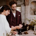 Braut und Bräutigam stehen in einer rustikalen Kirchenumgebung am Altar und fangen so die Essenz der Hochzeitsplanung auf wunderbare Weise ein. Die Braut, in einem weißen Kleid und Schleier, zündet zusammen mit ihrem Bräutigam, der einen dunklen Anzug mit Fliege trägt, eine Kerze an. Kerzen und Blumenarrangements schmücken elegant den Altar.
