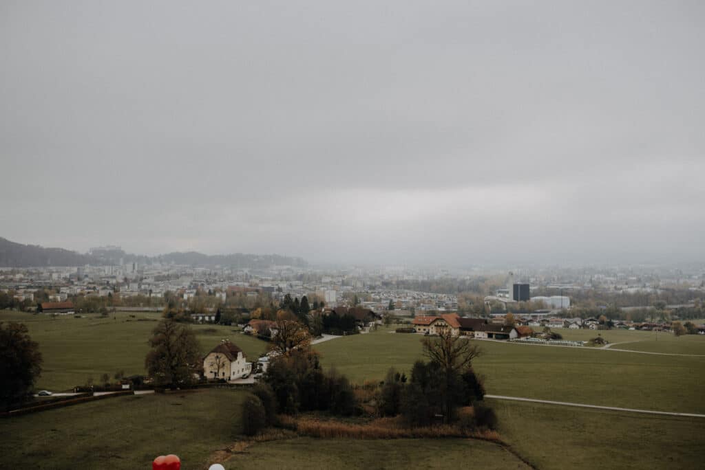 Luftaufnahme einer nebligen Landschaft mit grünen Feldern, vereinzelten Bäumen und wenigen Häusern, perfekt für eine verträumte Hochzeitslocation. In der Ferne taucht unter dem wolkigen Himmel die Skyline einer Stadt auf und bietet eine inspirierende Kulisse für elegante Hochzeitsplanung.
