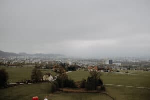 Luftaufnahme einer nebligen Landschaft mit grünen Feldern, vereinzelten Bäumen und wenigen Häusern, perfekt für eine verträumte Hochzeitslocation. In der Ferne taucht unter dem wolkigen Himmel die Skyline einer Stadt auf und bietet eine inspirierende Kulisse für elegante Hochzeitsplanung.