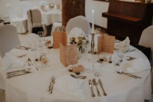 Ein runder Esstisch, elegant gedeckt für eine Hochzeit, mit weißem Tischtuch, gefalteten Servietten, Gläsern, Silberbesteck und braunen Menükarten. Eine große Kerze und ein kleines Blumengesteck werten das anspruchsvolle Dekor des Raumes auf, perfekt für Hochzeiten. Ein Klavier ziert den Hintergrund.