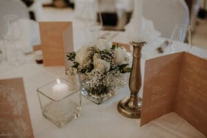Ein gedeckter Hochzeitstisch mit weißer Tischdecke besteht aus einer Kerze in einem Glashalter, einem Blumengesteck aus weißen Rosen und Schleierkraut, einem silbernen Kerzenhalter und braun bedruckten Menüs. Gläser und Geschirr sind ordentlich angeordnet.