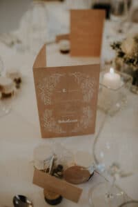 Ein Foto einer Hochzeitstafel mit einer braunen Menükarte mit weißen Blumenmustern. Auf dem Tisch stehen Gläser, eine brennende Kerze, Gläser mit Schnur und kleine Topfpflanzen, die eine elegante und gemütliche Atmosphäre schaffen, die perfekt für die Planung der idealen Hochzeitsfeier ist.