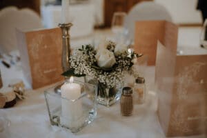 Auf einem Hochzeitstisch stehen eine weiße Kerze in einem Glashalter, ein Blumenarrangement mit weißen Rosen und Schleierkraut sowie Menükarten. Salz- und Pfefferstreuer stehen in der Nähe auf der weißen Tischdecke, elegant arrangiert für Ihre besondere Hochzeitsfeier.