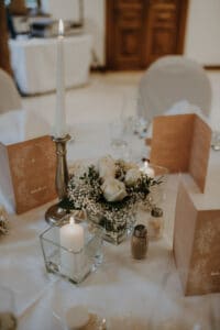 Auf einem dekorierten Hochzeitstisch stehen ein silberner Kerzenständer mit einer brennenden Stabkerze, eine Glasvase mit weißen Rosen und Schleierkraut sowie elegante braune Menükarten. Salz- und Pfefferstreuer sowie eine kleinere quadratische Kerze runden das Ambiente perfekt ab, wenn Sie schon tief in der Hochzeitsplanung stecken.
