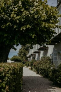 Ein von üppigen Büschen und blühenden Pflanzen gesäumter Weg führt zu einer Reihe weißer Häuser mit Balkonen – perfekt als Hochzeitskulisse. Ein großer Baum mit dichtem Laub wirft Schatten auf den Weg und schafft eine ruhige, sonnenbeschienene Gartenszene, die sich ideal eignet, um unvergessliche Hochzeitserinnerungen festzuhalten.