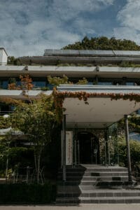 Ein modernes Gebäude mit Glaseingang und üppigem Grün an der Fassade, perfekt für eine Hochzeit. Solarmodule säumen das Dach und verleihen nachhaltigen Charme, während Sitzgelegenheiten im Freien einen idealen Platz für die Hochzeitsplanung unter dem teilweise bewölkten Himmel bieten.