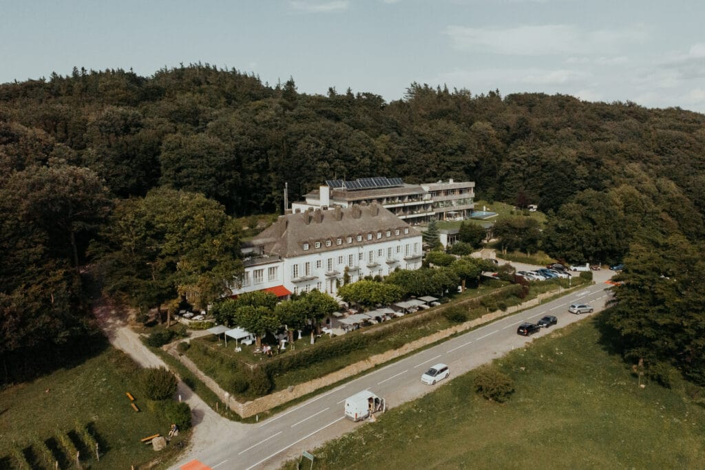 Eine Luftaufnahme zeigt ein großes, weißes, mehrstöckiges Hotel, eingebettet in eine üppige, bewaldete Gegend, perfekt für Hochzeitsplanung. Im Vordergrund ist eine Straße mit geparkten Fahrzeugen zu sehen, während Sitzplätze im Freien vorn auf einen idealen Ort für eine Hochzeit hindeuten.