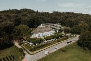 Eine Luftaufnahme zeigt ein großes, weißes, mehrstöckiges Hotel, eingebettet in eine üppige, bewaldete Gegend, perfekt für Hochzeitsplanung. Im Vordergrund ist eine Straße mit geparkten Fahrzeugen zu sehen, während Sitzplätze im Freien vorn auf einen idealen Ort für eine Hochzeit hindeuten.
