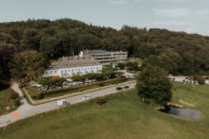 Luftaufnahme eines großen Hotels in einem Waldgebiet, perfekt für eine Traumhochzeit. Umgeben von Grün bietet es sowohl moderne als auch traditionelle Architektur. Mehrere Autos sind in der Nähe geparkt und deuten auf die Vorbereitungen für die Hochzeit neben dem malerischen Teich hin.