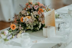 Ein wunderschön arrangierter Tischschmuck für die Hochzeit mit einem Strauß bunter Blumen, darunter Rosen und Gänseblümchen. Daneben befinden sich eine Menükarte, eine kleine Geschenkbox mit Schleife und klare Gläser, alles auf einer weißen Tischdecke – perfekt für Ihre Hochzeitsfeier.