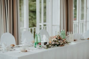 Ein langer, eleganter Esstisch ist für eine Hochzeitsveranstaltung gedeckt, mit weißem Tuch bedeckt und mit mehreren grünen Glasflaschen, Weingläsern, einem Blumengesteck und Menükarten geschmückt. Im Hintergrund sind große Fenster mit weichen, neutralen Vorhängen zu sehen. Perfekt für eine reibungslose Hochzeitsplanung.