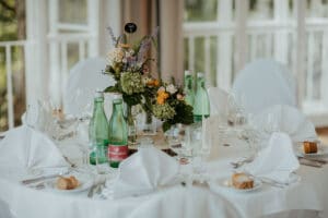 Ein runder Tisch ist elegant für eine Hochzeit gedeckt, mit weißen Tischdecken, Servietten, Glaswaren und grünen Glasflaschen mit Blumendekorationen. Die Stühle sind mit weißen Vorhängen bedeckt und der Veranstaltungsort ist hell, mit natürlichem Licht, das durch große Fenster hereinströmt – perfekt für Ihre Hochzeitsplanung.