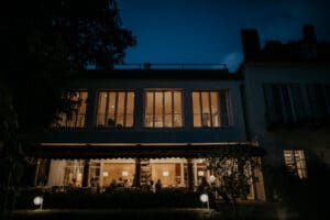 Ein großes Gebäude mit beleuchteten Fenstern bietet nachts die perfekte Kulisse für eine Hochzeit. Man sieht Menschen drinnen, die in einem warm beleuchteten Innenraum sitzen und stehen und vielleicht Einzelheiten der Hochzeit besprechen. Das Äußere ist dunkel, im Vordergrund sind teilweise Bäume und Büsche zu sehen.