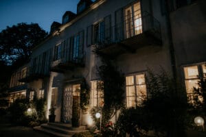Ein hohes Gebäude in der Abenddämmerung mit mehreren beleuchteten Fenstern und kleinen Balkonen, perfekt für alle, die von einer Märchenhochzeit träumen. Das Gebäude ist von Bäumen und Pflanzen umgeben, während kugelförmige Lichter den Weg in der Nähe des Eingangs beleuchten. Der tiefblaue Himmel signalisiert die Magie des frühen Abends.