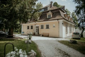 Ein charmantes gelbes Häuschen mit steilem Dach und Dachfenstern liegt inmitten üppiger Vegetation und eignet sich perfekt für eine intime Hochzeitsplanung. Ein Kiesweg führt zum Eingang, der von Bäumen und Blumen umgeben ist und eine romantische Kulisse bietet. In der Nähe steht ein kleines Auto für alle Hochzeitsbedürfnisse bereit.