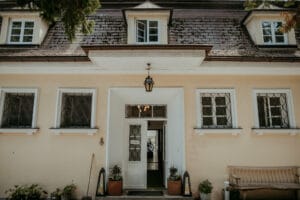Ein charmantes Haus mit schrägem Dach, perfekt für eine intime Hochzeitsplanung, mit einer offenen Eingangstür mit Glaspaneelen, flankiert von zwei kleinen Fenstern. Darüber hängt eine Verandalampe und eine Bank mit Topfpflanzen verleiht dieser idealen Hochzeitskulisse eine einladende Note.