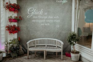 Eine gemütliche Terrasse mit einer Holzbank vor einer Wand mit dem Text: „Glück ist das Einzige das sich verdoppelt, wenn man es teilt.“ Perfekt für intime Heiratsmomente: Neben der Bank hängen lebendige Blumendekorationen in Pflanzgefäßen.