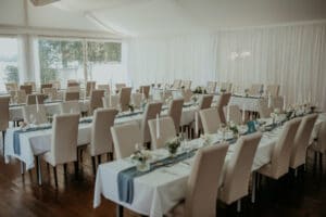 Ein perfekter Ort für Ihre Hochzeitsfeier mit Reihen von Tischen und Stühlen. Die Tische sind elegant mit weißen Tischdecken, blauen Läufern und Blumendekorationen gedeckt. Natürliches Licht fällt durch große Fenster in den Raum und erhellt das Dekor für Ihren besonderen Tag wunderschön.