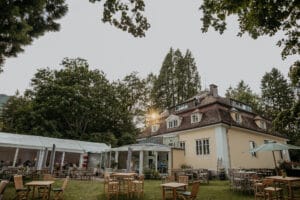 Eine charmante Villa mit großen Fenstern liegt inmitten üppiger Bäume und eines Gartens, perfekt für eine Hochzeitsfeier. Die Sonne scheint durch das Laub und beleuchtet die Sitzgelegenheiten im Freien mit Holztischen und -stühlen. Ein weißes Zelt verstärkt die ruhige Atmosphäre, ideal für Hochzeiten.