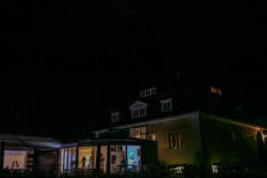 Ein Haus bei Nacht mit beleuchteten Fenstern zeigt die Silhouetten von Menschen im Inneren, die an einer fröhlichen Hochzeitsfeier teilnehmen. Der dunkle Himmel und das schwach beleuchtete Äußere verleihen dieser intimen Hochzeitsszene eine heitere, geheimnisvolle Atmosphäre.