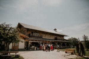 Ein rustikales Holzgebäude unter klarem Himmel beherbergt eine Gruppe von Menschen in traditioneller Kleidung, die eine Hochzeit feiern. Der Außenbereich, der sich perfekt für die Hochzeitsplanung eignet, bietet Bänke und Grünflächen und schafft eine malerische und ruhige Umgebung.