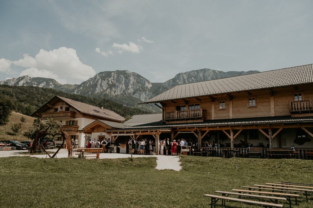 Eine wunderschöne Berghütte mit Holzfassade ist von Grasfeldern und majestätischen Bergen umgeben und bietet eine idyllische Kulisse für eine zauberhafte Hochzeit. Die Leute haben sich draußen versammelt, einige besprechen die Hochzeitsplanung, während der teilweise bewölkte Himmel zur heiteren Atmosphäre beiträgt.