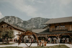 Eine rustikale Berghütte dient als charmanter Veranstaltungsort, an dem sich Menschen in traditioneller Kleidung zu einer herzlichen Hochzeit versammeln. Im Vordergrund steht eine herzförmige Holzskulptur, die die Liebe inmitten hoch aufragender Berge unter einem teilweise bewölkten Himmel symbolisiert. Perfekt für alle, die eine einzigartige Hochzeitsplanung suchen.