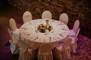 Ein runder Tisch, elegant gedeckt für eine formelle Hochzeit, bedeckt mit einer weißen Tischdecke und umgeben von sechs Stühlen mit weißen Hussen. Das Mittelstück aus Kerzen und Blumen schafft eine einladende und gehobene Atmosphäre vor der rustikalen Backsteinwand – perfekt für eine durchdachte Hochzeitsplanung.