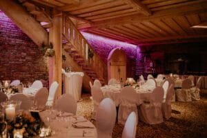Ein warm beleuchteter Hochzeitsempfang in einem rustikalen Raum mit Backsteinmauern und Holzbalken. Die Tische sind elegant mit weißen Tischdecken, Stühlen mit Hussen, Kerzen und Blumenarrangements gedeckt. Eine Holztreppe und stimmungsvolles violettes Licht verleihen der reizvollen Hochzeitsatmosphäre Charme.