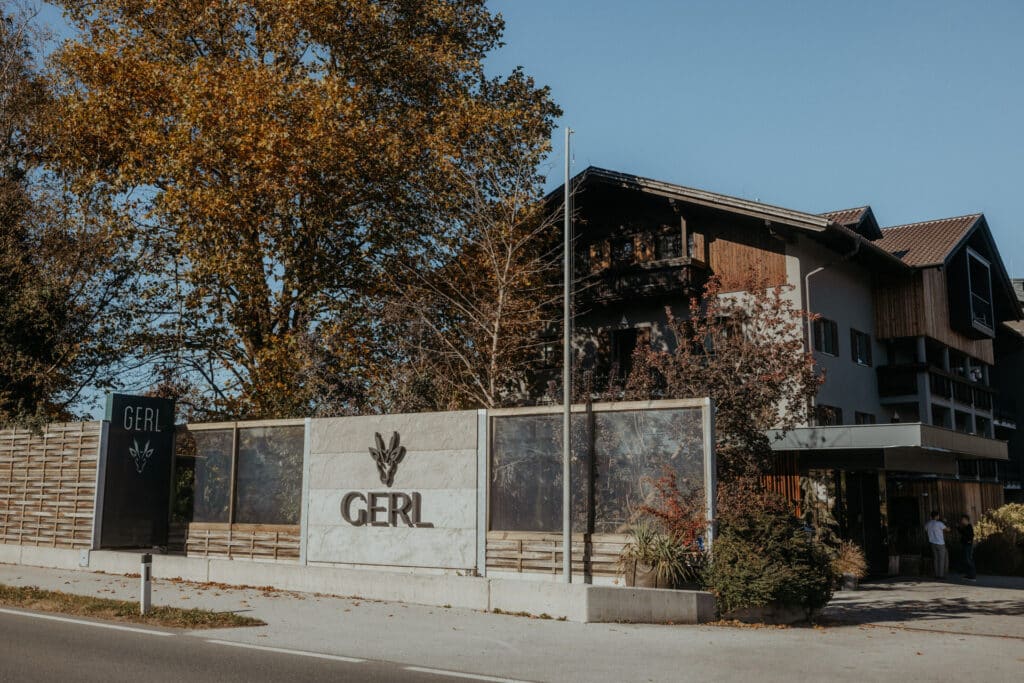Ein modernes Gebäude mit der Aufschrift "GERL" steht an einer von Bäumen gesäumten Straße unter einem strahlend blauen Himmel. Das Gebäude besteht aus Holz- und Steinelementen, und der Bereich ist mit Sträuchern und Bänken begrünt.