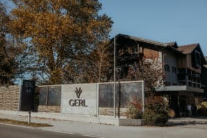 Ein modernes Gebäude mit der Aufschrift "GERL" steht an einer von Bäumen gesäumten Straße unter einem strahlend blauen Himmel. Das Gebäude besteht aus Holz- und Steinelementen, und der Bereich ist mit Sträuchern und Bänken begrünt.