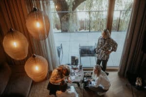 Vier Frauen befinden sich in einem gemütlichen Raum mit warmer Beleuchtung und großen Fenstern. Zwei sitzen auf dem Boden und schminken eine dritte Frau, während eine andere in der Nähe steht und zusieht. Draußen sind durch die Glastüren eine Terrasse und ein Baum zu sehen.