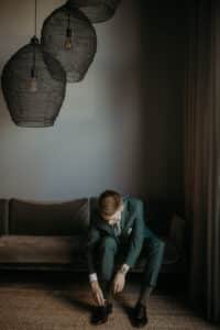 Ein Mann in einem blaugrünen Anzug sitzt auf einer grauen Couch in einem schwach beleuchteten Raum und beugt sich vor, um seine braunen Schuhe zu binden. Über ihm hängen drei große Pendelleuchten aus Draht, und rechts ist ein Vorhang zu sehen.