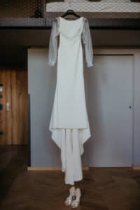 Ein langes weißes Hochzeitskleid mit durchsichtigen langen Ärmeln hängt auf einem Kleiderbügel. Darunter steht ein Paar goldene Stöckelschuhe auf einem Chevron-Holzboden in einem modernen Raum mit neutralen Wänden.