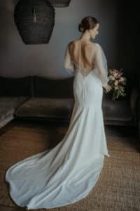 Eine Braut steht drinnen auf einem gewebten Teppich und trägt ein elegantes weißes Kleid mit einer langen Schleppe und durchsichtigen langen Ärmeln. Das Kleid hat einen tiefen, offenen Rücken mit zarten Knöpfen und Spitzendetails. Sie hält einen Blumenstrauß in der Hand und blickt weg.