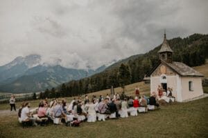 Eine kleine Hochzeitszeremonie im Freien findet neben einer malerischen Kapelle in einer Berglandschaft statt. Die Gäste sitzen auf Bänken und beobachten das Paar am Altar. Der Himmel ist bedeckt, im Hintergrund sind Wälder und Berge zu sehen.