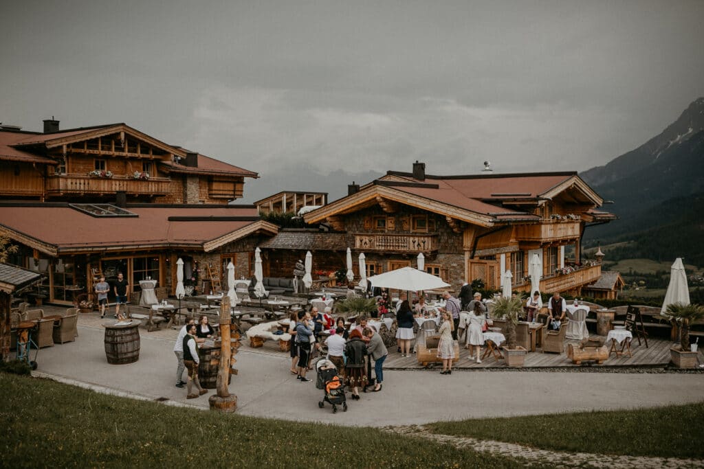 Eine rustikale Holzhütte, umgeben von Menschen, die ein Treffen im Freien genießen, bietet die perfekte Kulisse für eine gemütliche Hochzeit. Eingebettet in eine Berglandschaft unter einem bewölkten Himmel verstärken Tische, Stühle und üppiges Grün die gesellige Atmosphäre dieses idealen Ortes für Hochzeitsplanung.