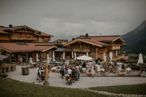 Eine rustikale Holzhütte, umgeben von Menschen, die ein Treffen im Freien genießen, bietet die perfekte Kulisse für eine gemütliche Hochzeit. Eingebettet in eine Berglandschaft unter einem bewölkten Himmel verstärken Tische, Stühle und üppiges Grün die gesellige Atmosphäre dieses idealen Ortes für Hochzeitsplanung.