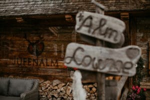 Außenansicht einer rustikalen Holzhütte mit dem Schild „Alm Lounge“ im Mittelpunkt. An einer Wand ist „Jufenalm“ mit einem Logo aus Hirschgeweih zu sehen, perfekt für intime Hochzeitsfeiern. Darunter sind Holzscheite gestapelt und links steht ein graues Korbsofa, das eine gemütliche, einladende Atmosphäre schafft, ideal für die Hochzeitsplanung.