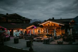 Ein gemütliches, beleuchtetes Holzchalet in einer Berggegend bietet in der Abenddämmerung eine perfekte Kulisse für intime Zusammenkünfte oder sogar zum Heiraten. Der Himmel ist bewölkt und warme Lichter heben das Gebäude hervor. Sitzgelegenheiten im Freien mit Tischen und Fässern schaffen eine einladende Atmosphäre, die sich ideal für jeden besonderen Anlass eignet.
