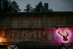 Ein Holzgebäude mit der Aufschrift „Jufenalm“ ist schwach beleuchtet, perfekt für eine romantische Hochzeit. Ein rosafarbener Scheinwerfer beleuchtet die Silhouette eines Hirschgeweihs an der Wand. Hohe Kiefern heben sich gegen den dunkler werdenden Himmel ab und bilden eine zauberhafte Kulisse, die sich ideal für Heiratspläne eignet.