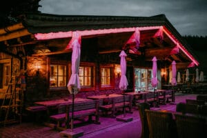 Eine gemütliche Restaurantterrasse im Freien in der Abenddämmerung, perfekt für die Hochzeitsplanung, mit Holzbänken und Tischen unter geschlossenen Sonnenschirmen. Warmes Licht leuchtet aus den Fenstern, während rote Neonlichter das rustikale Gebäude umrahmen. Der bedeckte Himmel verleiht diesem idealen Ort zum Heiraten eine romantische Note.