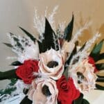 Ein bezaubernder Blumenstrauß für Ihre Hochzeitsplanung, bestehend aus cremefarbenen und roten Blüten mit dunkler Mitte, akzentuiert durch dünnes weißes getrocknetes Laub und dunkelgrüne Blätter, vor einem schlichten Hintergrund.