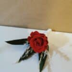 Eine einzelne rote Rose, Symbol der Liebe und Verbundenheit, mit grünen Blättern und weißen Akzentblüten liegt auf einer weißen Fläche vor einem braunen Hintergrund und wirft einen sanften Schatten – eine perfekte Note für die Hochzeitsplanung.