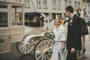 Braut und Bräutigam stehen neben einer weißen Pferdekutsche auf einer Stadtstraße. Die Braut hält einen Blumenstrauß und lächelt. Sie trägt eine weiße Pelzstola über ihrem Hochzeitskleid. Der Bräutigam, in einem dunklen Anzug, blickt sie mit einem sanften Lächeln an und verkörpert damit die Freude ihrer Hochzeit.
