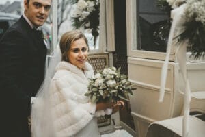 Braut und Bräutigam lächeln neben einer mit Blumen geschmückten Oldtimerkutsche und fangen damit die Essenz ihrer Hochzeit ein. Die Braut, in einem weißen Pelzschal und mit einem Strauß weißer Blumen in der Hand, steht anmutig neben dem Bräutigam in seinem schwarzen Anzug und verkörpert elegante Hochzeitsplanung.
