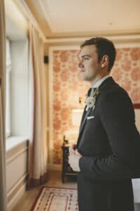 Ein Mann im Anzug und mit Boutonnière steht drinnen neben einem Fenster und scheint Teil einer eleganten Hochzeit zu sein. Die gemusterte Tapete und der Teppich des Raums schaffen eine warme Atmosphäre für die Hochzeitsszene. Er blickt ruhig nach vorne, während natürliches Licht diesen Moment der Hochzeitsplanung erhellt.