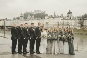 Braut und Bräutigam stehen mit ihrer Hochzeitsgesellschaft an einem Uferweg und verkörpern damit die Schönheit von Hochzeit. Die Brautjungfern tragen graue Kleider und halten Blumensträuße, während die Trauzeugen schwarze Anzüge tragen. Eine historische Stadtlandschaft und ein Schloss bilden eine atemberaubende Kulisse unter dem bewölkten Himmel.