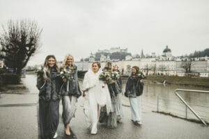 Eine Gruppe von Frauen in eleganten Kleidern und Pelzschals, vermutlich Teil einer Hochzeitsfeier, geht an einem bewölkten Tag an einem Fluss entlang. Sie halten Blumensträuße in den Händen und lächeln. Im Hintergrund ist eine Stadtlandschaft mit historischen Gebäuden und einem Schloss auf einem Hügel zu sehen.