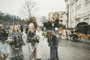Eine fröhliche Hochzeitsgesellschaft geht in eleganten Kleidern und Schals eine Straße entlang und plaudert fröhlich über ihre Hochzeit. Hinter ihnen wartet in der malerischen Stadt mit ihrer klassischen Architektur eine Pferdekutsche, die die perfekte Kulisse für ihre Hochzeitsfeier bietet.